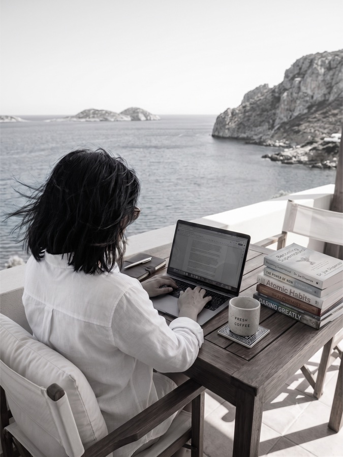 The author, seated at an outdoor desk overlooking the sea, writing on a laptop beside a stack of well-worn books.